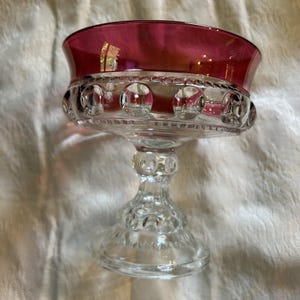 May include: A clear glass dessert dish with a ruby red rim. The bowl has a decorative pattern of circles around the rim. The stem and base are also clear glass with detailed designs. This vintage piece is ideal for serving desserts.