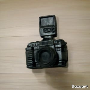 May include: A black camera with a flash attachment. The camera has a lens, buttons, and dials. The camera is sitting on a light brown wooden surface. The word "Bazaart" is in the bottom right corner.