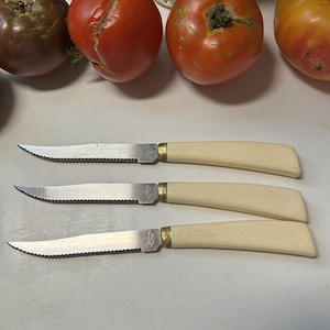 May include: Three steak knives with serrated stainless steel blades, brass-colored metal bolsters, and off-white plastic handles. The knives are arranged horizontally, each featuring a pointed tip and a metallic finish on the blade.