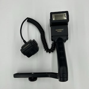 May include: Black Quantaray camera flash with a handle and a coiled cord. The flash unit has a rectangular head and a bracket for mounting. The text "QUANTARAY" is visible on the flash and the remote unit.