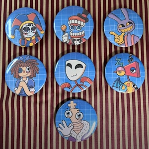May include: A collection of seven round, blue pinback buttons featuring cartoon characters. The characters include a jester, a toothy figure in a tuxedo, a purple rabbit, a rag doll, a white mask, a robot, and a king. The buttons are arranged on a striped background.