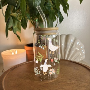May include: A clear glass tumbler with a wooden lid and a glass straw. The tumbler is decorated with illustrations of white geese, flowers, and strawberries. The tumbler is sitting on a wooden tray.