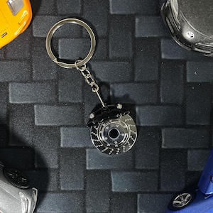 Op de afbeelding: Een zilverkleurige sleutelhanger met een remschijf en een zwarte remklauw. De sleutelhanger is bevestigd aan een zilveren ring en ketting. Speelgoedauto's in geel, zwart, zilver en blauw omringen de sleutelhanger.