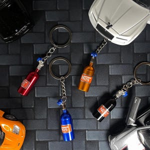 May include: Keychains featuring miniature NOS bottles in red, orange, blue, and black. Each keychain has a silver chain and ring. The bottles have the word "NOS" printed on them. Toy cars are in the background.