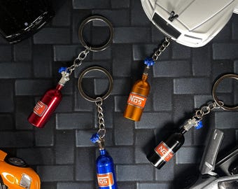 May include: Keychains featuring miniature NOS bottles in red, orange, blue, and black. Each keychain has a silver chain and ring. The bottles have the word "NOS" printed on them. Toy cars are in the background.