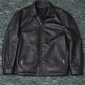 Könnte beinhalten: Schwarze Lederjacke mit Reißverschluss und zwei Seitentaschen. Die Jacke hat einen Kragen und Knöpfe. Sie wird auf einer strukturierten, dunkelgrünen Oberfläche präsentiert.