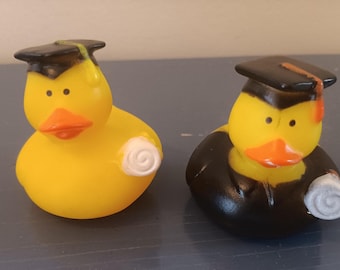 May include: Two yellow rubber ducks wearing graduation caps and holding scrolls. One duck is entirely yellow, while the other has a black body. Both have orange beaks and are on a dark surface.