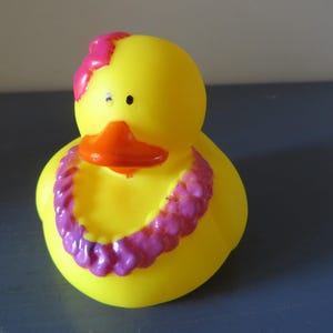 Op de afbeelding: Een gele rubberen eend met een oranje snavel en een roze bloem op zijn hoofd. De eend is versierd met een paarse en roze lei-ketting, wat een speels en tropisch thema cre&euml;ert.