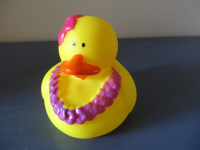 Op de afbeelding: Een gele rubberen eend met een oranje snavel en een roze bloem op zijn hoofd. De eend is versierd met een paarse en roze lei-ketting, wat een speels en tropisch thema cre&euml;ert.