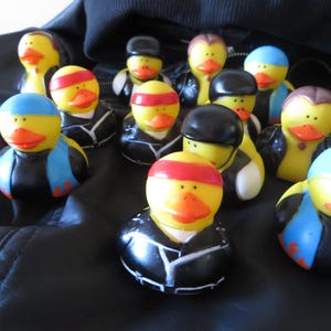 May include: A group of yellow rubber duck figurines, each with a different outfit. Some ducks wear black jackets, others have headbands or hats. The ducks have orange beaks and are on a black surface.