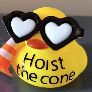 Op de afbeelding: Een gele rubberen eend met een zwarte hartvormige zonnebril met witte glazen en een kleine oranje en witte verkeerskegel. De tekst "Hoist the cone" staat op het lichaam van de eend.