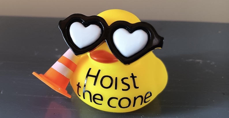 Op de afbeelding: Een gele rubberen eend met een zwarte hartvormige zonnebril met witte glazen en een kleine oranje en witte verkeerskegel. De tekst "Hoist the cone" staat op het lichaam van de eend.