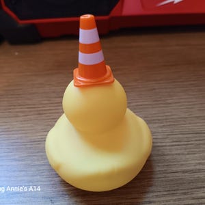 Op de afbeelding: Een gele rubberen eend met een kleine oranje en wit gestreepte verkeerskegel op zijn hoofd. De eend heeft een klassieke, afgeronde vorm en staat op een houten oppervlak.