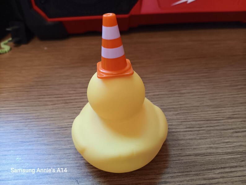 Op de afbeelding: Een gele rubberen eend met een kleine oranje en wit gestreepte verkeerskegel op zijn hoofd. De eend heeft een klassieke, afgeronde vorm en staat op een houten oppervlak.