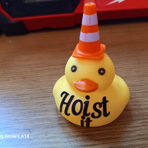 Op de afbeelding: Een gele rubberen eend met zwarte ogen en een oranje snavel, met een oranje en witte verkeerskegelhoed. De tekst "Hoist it" staat op de borst van de eend.