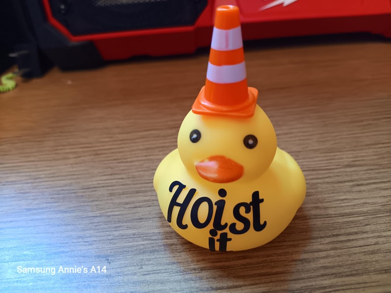 Op de afbeelding: Een gele rubberen eend met zwarte ogen en een oranje snavel, met een oranje en witte verkeerskegelhoed. De tekst "Hoist it" staat op de borst van de eend.