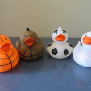 May include: Four rubber duck toys, each decorated with a different sport. One is orange and black with a basketball design, one is brown with a basketball design, one is white with a soccer ball design, and one is white with a baseball design.