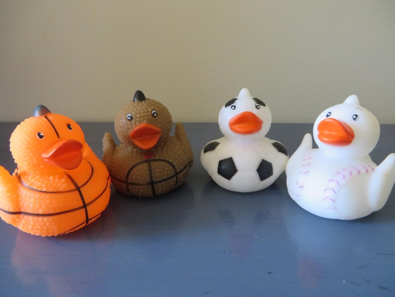 May include: Four rubber duck toys, each decorated with a different sport. One is orange and black with a basketball design, one is brown with a basketball design, one is white with a soccer ball design, and one is white with a baseball design.