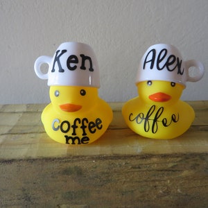 Puede incluir: Dos patos de goma amarillos con tazas de café blancas en la cabeza. Las tazas tienen los nombres "Ken" y "Alex" escritos en ellas. Los patos tienen las palabras "coffee me" escritas en sus pechos.