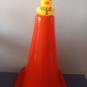 Op de afbeelding: Een feloranje verkeerskegel met een gele eendenopzet. De eend draagt een gestreepte oranje en witte hoed en heeft de woorden "Hoist it" gedrukt op een gele basis.