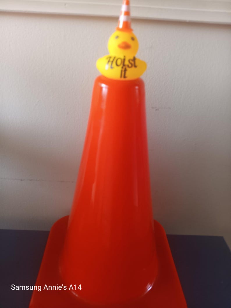 Op de afbeelding: Een feloranje verkeerskegel met een gele eendenopzet. De eend draagt een gestreepte oranje en witte hoed en heeft de woorden "Hoist it" gedrukt op een gele basis.
