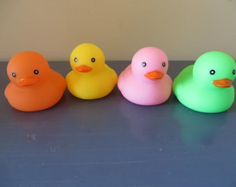 May include: Four colorful rubber duck toys in a row. The ducks are orange, yellow, pink, and green, each with black eyes and orange beaks. They are sitting on a dark blue surface.