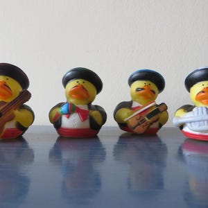 Könnte beinhalten: Vier gelbe Gummienten, die als Mariachi-Musiker verkleidet sind. Jede Ente trägt einen schwarzen Hut und ein Kostüm mit roten und weißen Akzenten. Eine Ente hält eine Gitarre, eine andere eine Geige und eine dritte eine Trompete.