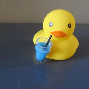 May include: A yellow rubber duck with a blue drink in a clear cup with a black straw. The duck has a black eye and a black beak.