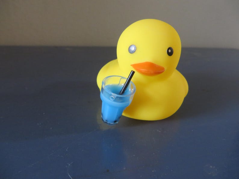 May include: A yellow rubber duck with a blue drink in a clear cup with a black straw. The duck has a black eye and a black beak.