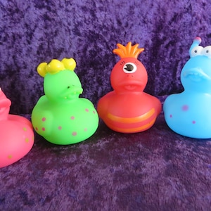 May include: Four colorful rubber duck toys in a row. The ducks are pink, green, red, and blue, each with unique features like spots, antennae, and a single eye. They are arranged on a purple, textured surface.