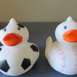 May include: Two white rubber duck toys. One duck is decorated with black and white soccer ball designs. The other duck is decorated with pink baseball stitching.
