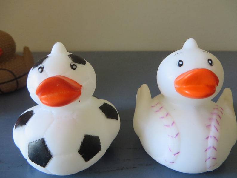 May include: Two white rubber duck toys. One duck is decorated with black and white soccer ball designs. The other duck is decorated with pink baseball stitching.