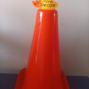 Op de afbeelding: Een feloranje verkeerskegel met een gele rubberen eend die een zonnebril draagt en een miniatuurkegel vasthoudt. Op de eend staat de tekst "Hoist the cone". De kegel staat op een donkerblauwe ondergrond.