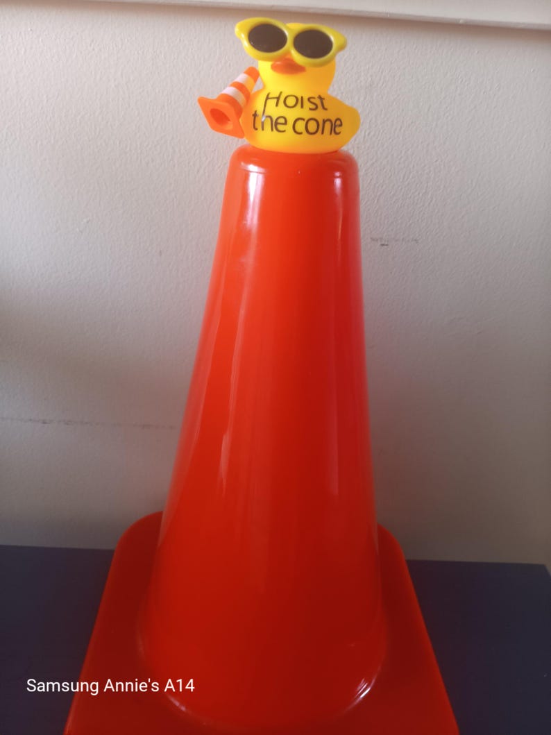 Op de afbeelding: Een feloranje verkeerskegel met een gele rubberen eend die een zonnebril draagt en een miniatuurkegel vasthoudt. Op de eend staat de tekst "Hoist the cone". De kegel staat op een donkerblauwe ondergrond.