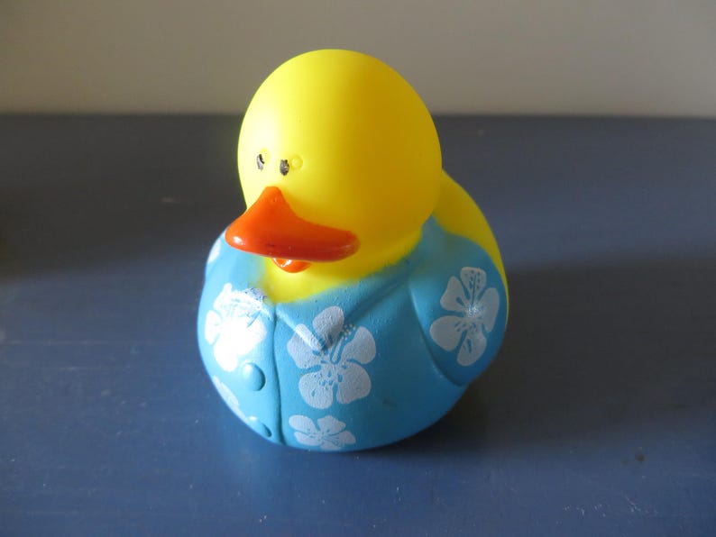 Op de afbeelding: Een gele rubberen eend met een oranje snavel en zwarte ogen. De eend draagt een blauw shirt met witte bloempatronen. Het speelgoed is ontworpen om op een eend te lijken.