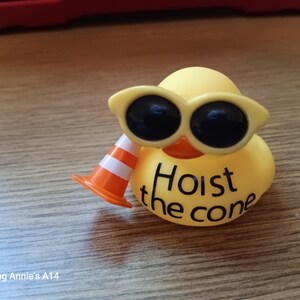 Op de afbeelding: Een gele rubberen eend met een gele zonnebril en een oranje en witte verkeerskegel. De tekst "Hoist the cone" staat op de borst van de eend. De eend staat op een houten oppervlak.
