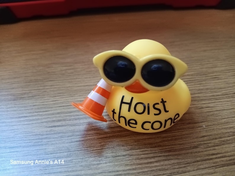 Op de afbeelding: Een gele rubberen eend met een gele zonnebril en een oranje en witte verkeerskegel. De tekst "Hoist the cone" staat op de borst van de eend. De eend staat op een houten oppervlak.