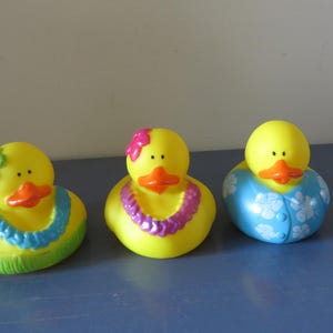 Op de afbeelding: Drie gele rubberen badeenden met oranje snavels en zwarte ogen. Elke eend is versierd met een ander tropisch thema, waaronder kransen en bloemmotieven. De eenden zijn gerangschikt op een blauw oppervlak.