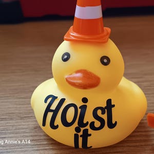 Op de afbeelding: Een gele rubberen eend met een oranje en witte verkeerskegel hoed. De eend heeft zwarte ogen en een oranje snavel. De woorden "Hoist it" staan in het zwart op het lichaam van de eend.