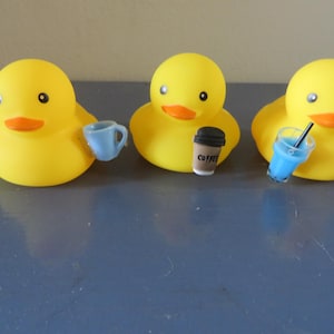 May include: Three yellow rubber duck toys, each holding a different beverage. The duck on the left is holding a blue cup, the middle duck is holding a brown coffee cup with the word "COFFEE" printed on it, and the duck on the right is holding a blue cup with a straw.