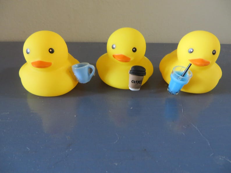 May include: Three yellow rubber duck toys, each holding a different beverage. The duck on the left is holding a blue cup, the middle duck is holding a brown coffee cup with the word "COFFEE" printed on it, and the duck on the right is holding a blue cup with a straw.