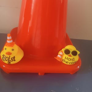 Op de afbeelding: Een feloranje verkeerskegel met een brede basis. Twee gele eendvormige ornamenten staan op de basis. E&eacute;n eend heeft een kleine verkeerskegelhoed en de tekst "Hoist it". De andere eend draagt een zonnebril en heeft de tekst "Hoist the cone".