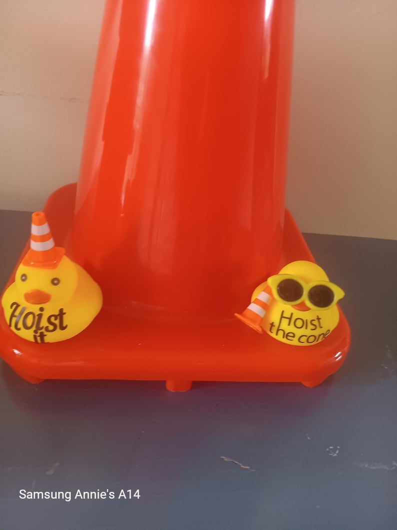 Op de afbeelding: Een feloranje verkeerskegel met een brede basis. Twee gele eendvormige ornamenten staan op de basis. E&eacute;n eend heeft een kleine verkeerskegelhoed en de tekst "Hoist it". De andere eend draagt een zonnebril en heeft de tekst "Hoist the cone".