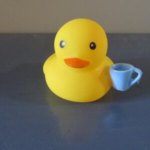 May include: A yellow rubber duck with a blue teacup in its beak. The duck has black eyes and an orange beak.
