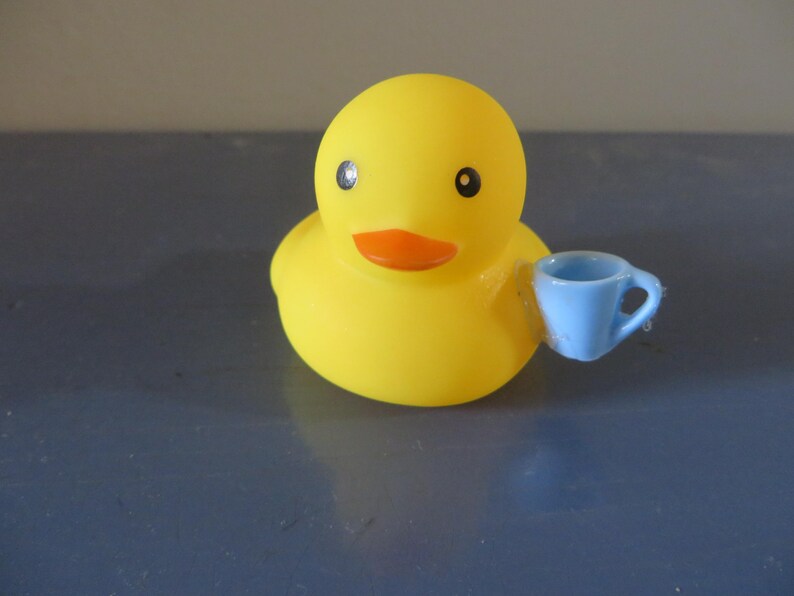 May include: A yellow rubber duck with a blue teacup in its beak. The duck has black eyes and an orange beak.