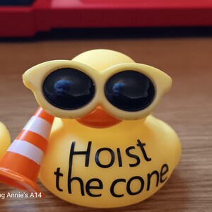 Op de afbeelding: Een gele rubberen eend met een zonnebril en een miniatuur oranje en witte verkeerskegel. De tekst "Hoist the cone" staat op het lichaam van de eend. Een leuk, nieuwigheid artikel.