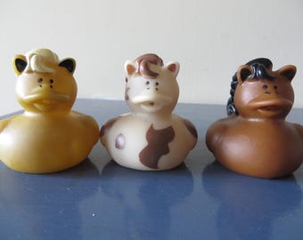 May include: Three rubber duck toys, each designed to resemble a different animal. The first is yellow with a duck bill and a horse-like mane. The second is white with brown spots and a brown mane. The third is brown with a black mane.