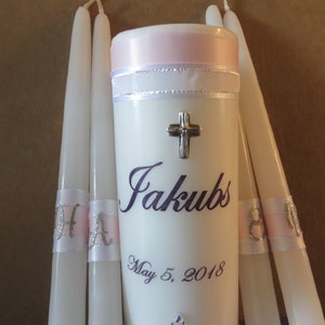May include: Set of four white baptism candles with pink and silver ribbon accents. The center candle has a silver cross and the name "Jakubs" and the date "May 5, 2018" printed in purple.