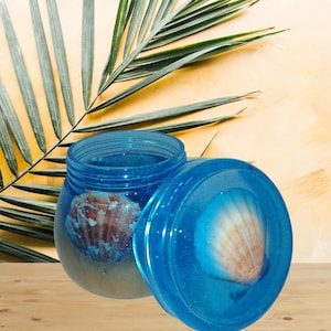 May include: A blue jar with a seashell inside, and a matching lid. The jar is open, revealing the seashell and a sandy base. The lid, also blue, has a seashell embedded in it. The background includes a palm leaf.