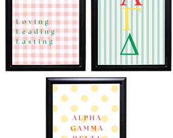May include: Three framed prints in black frames. One features a pink and white gingham pattern with the words "Loving, Leading, Lasting" in green. Another has green and white stripes with the letters A, Γ, and Δ in red, yellow, and green. The third print has yellow polka dots with the words "Alpha, Gamma, Delta" in red.